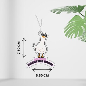 Könnte beinhalten: Ein weißer Gänse-Lufterfrischer mit einer Cartoon-Gans, die eine schwarze Sonnenbrille trägt. Der Text "AM ENDE KACKT DIE GANS!" ist in Schwarz und Pink gedruckt. Das Objekt misst 7,50 cm in der Höhe und 5,50 cm in der Breite, markiert durch Pfeile.