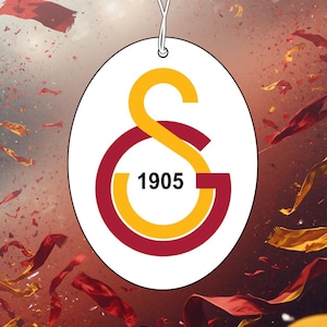 May include: An oval-shaped hanging air freshener featuring the Galatasaray sports club logo with a yellow S and a red G intertwined, centred with the black text 1905 on a white background, suspended by a thin white string for display.