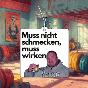 Pode incluir: Um ambientador quadrado com fundo branco, o texto preto "Muss nicht schmecken, muss wirken" e uma fotografia de uma pessoa com um capuz cinzento a segurar um copo de cerveja, suspenso por um fio branco contra um fundo de ginásio.