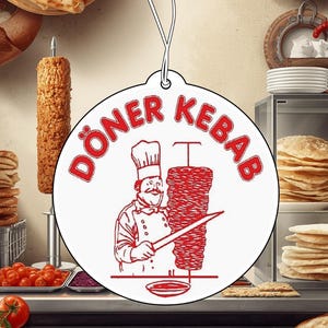Op de afbeelding: Een ronde witte hangende decoratie met een rode grafische illustratie van een kok die vlees snijdt van een verticale draaispit. De tekst DÖNER KEBAB staat in rode hoofdletters in een boog boven de kok en de draaispit gedrukt.
