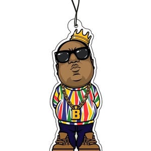 May include: A hanging air freshener featuring a cartoon illustration of a person wearing a gold crown, black sunglasses, and a multi-coloured striped sweater. The figure wears a gold chain with a letter B pendant, dark blue trousers, and tan boots.