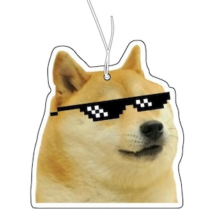 Op de afbeelding: Een uitgestanste luchtverfrisser met een beige Shiba Inu-hond die een zwarte, gepixelde zonnebril draagt. De hond heeft puntige oren en een lichte snuit. Aan de bovenkant is een wit touwtje bevestigd om hem op te hangen.