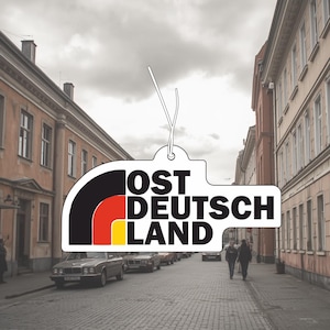 May include: A white, black, red, and yellow air freshener shaped like a stylized mountain logo with the text OST DEUTSCH LAND printed in bold black letters. The object features a small hole at the top with a thin white string attached for hanging.