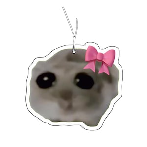 Op de afbeelding: Een hangend ornament met een afbeelding van een verdrietige, grijskleurige hamster met grote ogen en een felroze strik op het hoofd. Het ornament heeft een witte rand en bevat een dun wit touwtje aan de bovenkant om het op te hangen als decoratie.