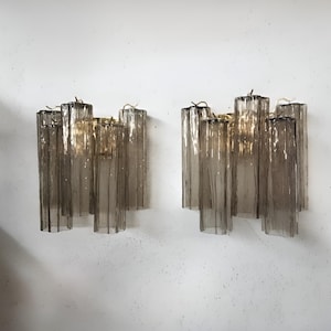 May include: Two matching wall sconces with a mid-century modern design. Each sconce features multiple vertical, rectangular glass rods in shades of gray and brown, mounted on a gold-toned metal base. The sconces are mounted on a white wall.