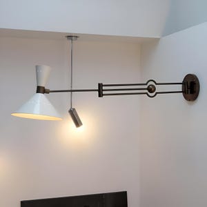 May include: A contemporary wall lamp with a white, cone-shaped shade and a small cylindrical light. The lamp has a dark metal arm and a circular wall mount. The design includes a pivoting arm and a hanging light fixture.