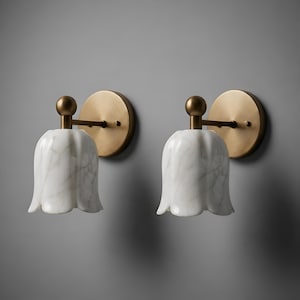 May include: Two matching wall sconces. Each features a white marble tulip-shaped shade, a brass arm, and a round brass backplate. The sconces are mounted on a gray wall, providing a soft, ambient light.