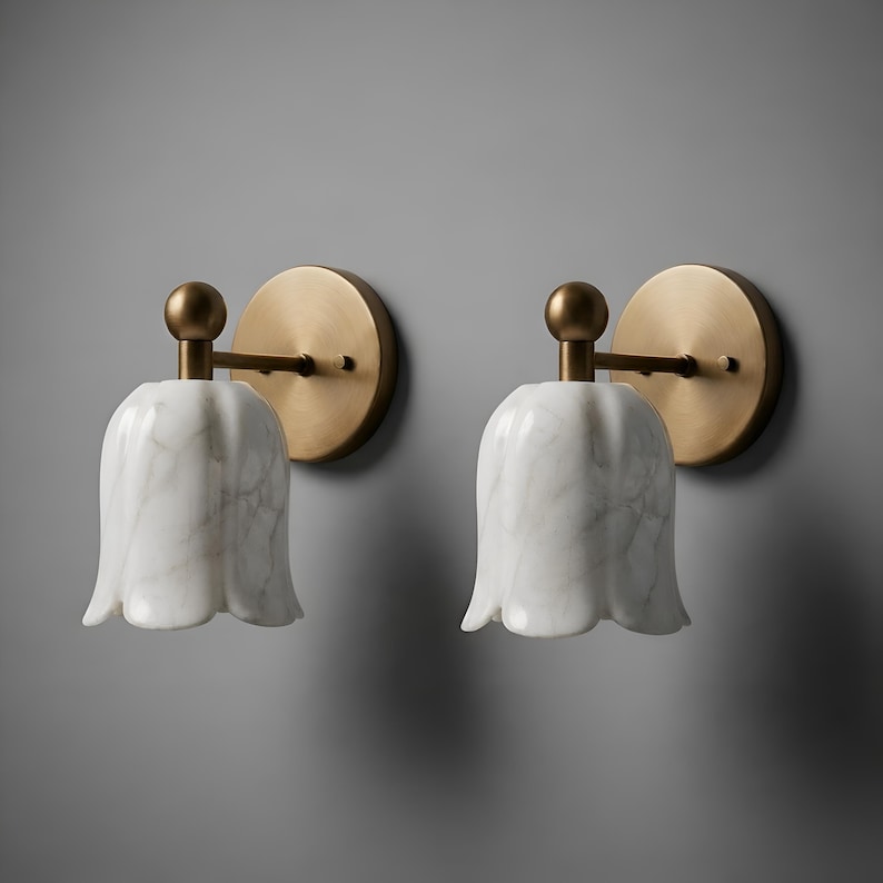 May include: Two matching wall sconces. Each features a white marble tulip-shaped shade, a brass arm, and a round brass backplate. The sconces are mounted on a gray wall, providing a soft, ambient light.