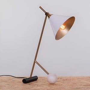 May include: A modern desk lamp with a white conical shade and a gold interior. The lamp has a slender, gold-colored frame and a black base with a white marble sphere. The light bulb is visible inside the shade.