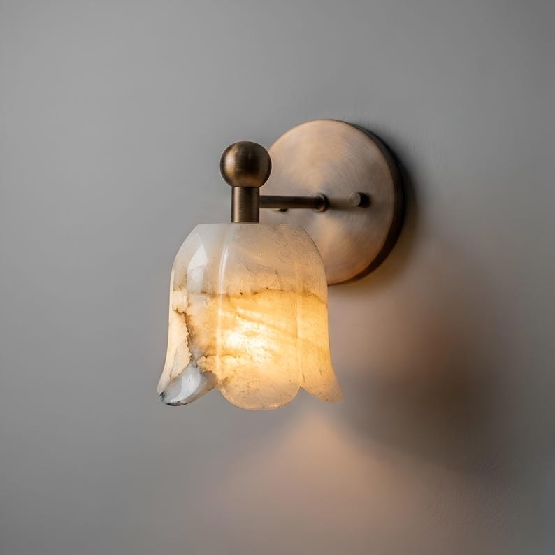 May include: A wall-mounted sconce with a tulip-shaped shade made of translucent white and tan stone. The sconce has a brass-colored metal base and arm, and a round backplate. The light is on, casting a warm glow.