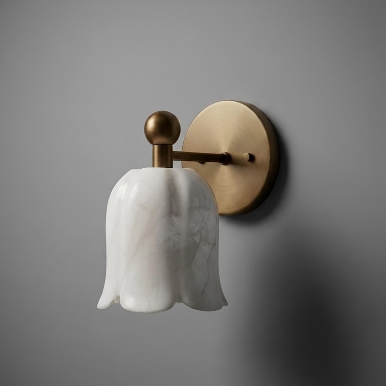 May include: A wall-mounted light fixture with a white, tulip-shaped shade with marble veining. The fixture has a brass-colored circular backplate and a brass arm. The overall design is elegant and decorative.
