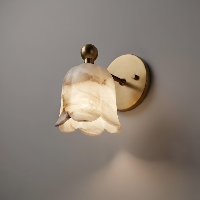 May include: A wall sconce with a tulip-shaped shade made of translucent, off-white stone with brown veining. The sconce has a brass-colored base and a small, round finial. The fixture is mounted on a round brass backplate.