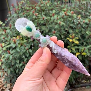 Puede incluir: Una daga de cristal púrpura y blanco tallada con detalles de piedras preciosas verdes. La daga presenta una parte superior redondeada con un cristal transparente y una piedra preciosa verde en el centro. El mango es de color púrpura claro y la hoja es de un púrpura más oscuro.