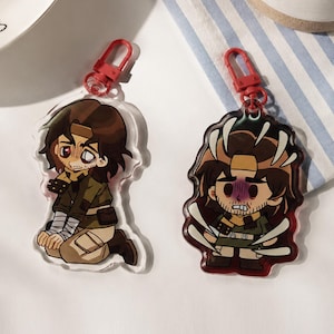 May include: Two acrylic keychains featuring a cartoon character with brown hair and a headband. The left keychain shows the character kneeling in a green shirt and tan pants. The right keychain shows the character standing with white spikes.