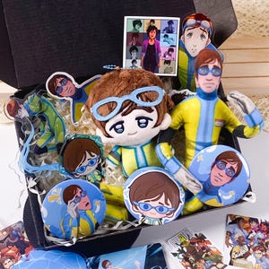 May include: A black gift box filled with merchandise featuring a character in a yellow and blue suit with goggles. Items include a plush doll, two circular buttons, acrylic charms, a square print, and stickers depicting the same character.