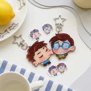 Könnte beinhalten: Zwei Acryl-Schlüsselanhänger mit Cartoon-Figuren und sternförmigen Verschlüssen, begleitet von zwei kleinen Aufklebern. Eine Figur hat braunes Haar und eine blaue Brille, die andere dunkles Haar und einen Bartansatz. Beide zeigen Gesichter.