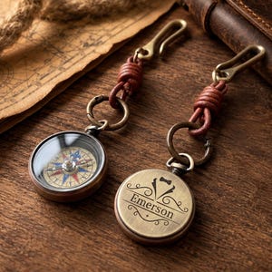 May include: Two round brass compasses with brown leather cord attachments and metal hooks. One compass shows a detailed navigation dial with blue and red accents, while the other features an engraved tuxedo icon and the name Emerson.
