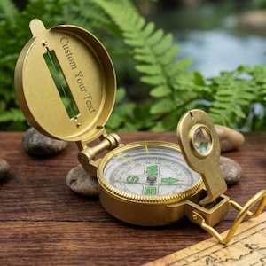 Peut inclure: Une boussole de poche en métal doré avec un couvercle ouvert portant le texte gravé Custom Your Text. La boussole présente un cadran blanc avec des marqueurs directionnels verts, une petite loupe sur bras articulé et un fil de visée.