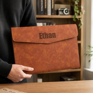 May include: A rectangular brown document folder with a textured finish and a flap closure. The name Ethan is engraved in a dark, sans-serif font on the center of the front flap. The folder has a slim profile and a smooth, consistent brown color.