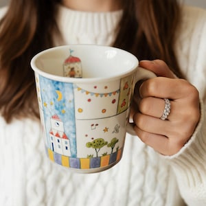 Puede incluir: Taza de cerámica blanca con un diseño ilustrado caprichoso. La taza presenta una colorida escena de pueblo con un cielo azul, estrellas y una luna creciente. El asa es blanca y el interior también es blanco. La taza es sostenida por una persona que lleva un suéter blanco.