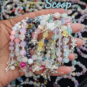 May include: A hand holds a variety of crystal bead bracelets in shades of pink, purple, blue, and yellow. The bracelets have silver charms and are displayed with the text "Bracelet Scoop" and "Get a random scoop of crystal bead bracelets."