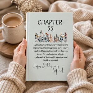 May include: A white book with the text "CHAPTER 55" and floral illustrations. The book's message reads "Celebrate everything you've become..." and includes a birthday message signed "Sophia!" A mug of coffee and a beige sweater are in the background.