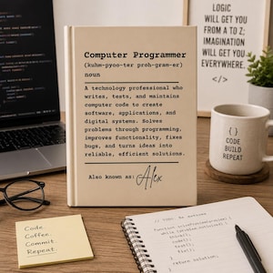 Puede incluir: Un libro de tapa dura color crema con una definición estilo diccionario de Computer Programmer impresa en texto negro, incluyendo el nombre Alex. El libro se muestra sobre un escritorio de madera junto a una computadora portátil, una taza y notas.