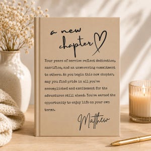 May include: A beige hardcover book featuring the black printed text 'a new chapter' with a heart symbol, followed by a paragraph honoring years of service and the name 'Matthew' at the bottom, presented in a clean, upright orientation.