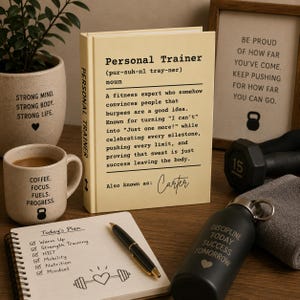 May include: A cream-colored book titled Personal Trainer with a definition, a beige mug with text and a kettlebell icon, a black water bottle with text, a spiral notebook with a workout plan, a black pen, a gray towel, and a framed sign with text.