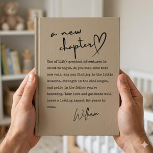 May include: A beige hardcover book with black text that reads: a new chapter, followed by a paragraph about fatherhood and the name William. The cover features a hand-drawn heart symbol next to the title, presented in a clean, minimalist style.