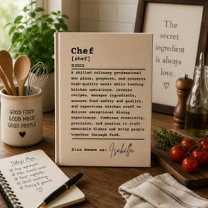 May include: A rectangular beige book featuring a dictionary-style definition of the noun Chef, including the text Also known as: Isabella. The book is displayed alongside a ceramic utensil holder, a spiral-bound notepad, and fresh tomatoes.