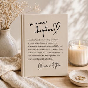 May include: A beige hardcover book featuring the black text 'a new chapter' with a heart symbol, followed by a printed paragraph and the names 'Olivia & Ethan'. The book cover has a clean, neutral design with a matte finish and clear typography.