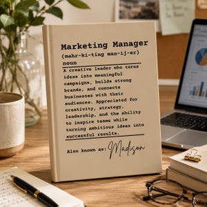 Può includere: Un libro color crema con testo nero che presenta una definizione in stile dizionario di Marketing Manager, inclusa la pronuncia fonetica, un paragrafo descrittivo e il testo personalizzato Also known as: Madison in carattere corsivo.
