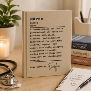 Könnte beinhalten: Ein beigefarbenes Hardcover-Buch mit einer wörterbuchartigen Definition des Wortes Nurse in schwarzem Druck. Die Definition beschreibt eine mitfühlende Pflegefachkraft, und unten steht der personalisierte Text Also known as: Evelyn.