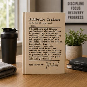 May include: A rectangular beige sign featuring black text that defines an Athletic Trainer as a healthcare and fitness professional. The text includes a phonetic pronunciation, a detailed definition, and the handwritten name Michael.