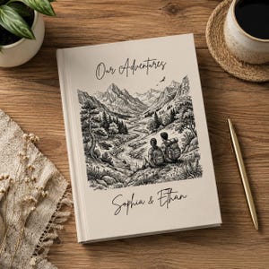 Könnte beinhalten: Ein cremefarbenes Notizbuch mit den Worten "Our Adventures" und den Namen "Sophia & Ethan" in Schriftform. Das Cover zeigt eine Schwarz-Weiß-Illustration einer Berglandschaft mit zwei Figuren. Ein goldener Stift und eine Tasse Kaffee liegen auf der Holzoberfläche.