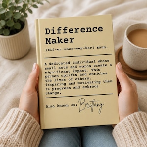 Può includere: Un libro giallo chiaro intitolato "Difference Maker" con una definizione e il nome "Brittany" in corsivo. Il libro è tenuto da una persona. Una tazza di caffè e una piccola pianta sono sullo sfondo.
