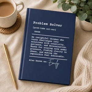 May include: A navy blue notebook with the words "Problem Solver" in white. The definition of a problem solver is printed below, along with the name "Emily". A gold pen and a cup of coffee are visible.