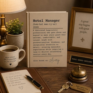 Può includere: Un libro beige intitolato Hotel Manager con una definizione in stile dizionario e il nome Avery stampato sulla copertina. Il libro è circondato da una tazza in ceramica, un blocco note, una penna, un campanello in ottone e una cornice.