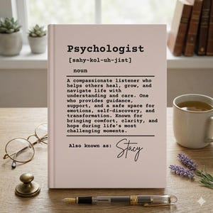 Puede incluir: Un libro rosa claro con la palabra "Psychologist" en letras negras en negrita. La definición de psicólogo está impresa debajo, junto con el nombre "Stacy" en cursiva. Una pluma, gafas y una taza de té están sobre la mesa.