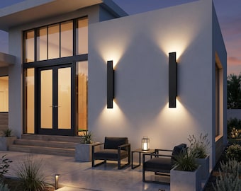 May include: Two rectangular black wall sconces mounted on a white exterior wall, emitting warm light upward and downward. The modern home features a double glass door entrance, two dark gray armchairs, a small side table, and concrete planters.
