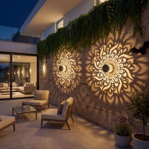 Op de afbeelding: Twee ronde buitenlampen voor aan de muur, gemonteerd op een stenen muur, die ingewikkelde, symmetrische, mandala-achtige lichtpatronen werpen op het gestructureerde oppervlak. De armaturen zijn donker met een warme, gloeiende lichtopbrengst.