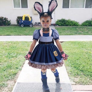 May include: A child dressed in a Zootopia Officer Judy Hopps costume. The outfit includes a blue dress, gloves with pink paw pads, bunny ears, and a bow tie. The costume is worn outdoors on a sunny day.