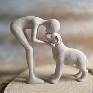 May include: A light beige sculpture of a person and a dog. The person is bent over, touching the dog's head. The dog is standing on all fours. The sculpture is on a wooden surface.
