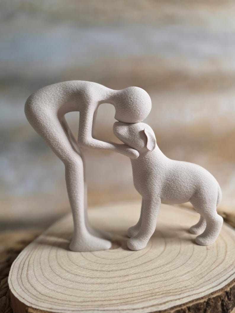 May include: A light beige sculpture of a person and a dog. The person is bent over, touching the dog's head. The dog is standing on all fours. The sculpture is on a wooden surface.