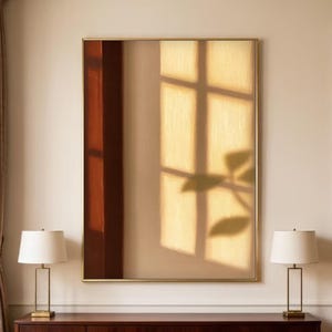 May include: A large, rectangular abstract painting with a gold frame. The artwork features a warm color palette with a window and plant shadow design. Two matching table lamps with white shades sit on a dark wood cabinet.