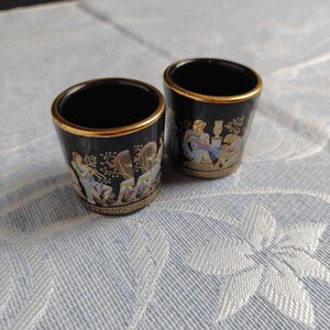 Könnte beinhalten: Zwei kleine, schwarze Schnapsgläser aus Keramik mit goldenen Rändern. Jedes Glas zeigt eine klassische Szene mit Figuren in farbigen Gewändern und Musikinstrumenten. Der Boden jedes Glases hat ein griechisches Schlüsselmuster.