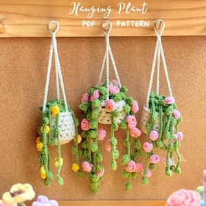 May include: Three crochet hanging plant patterns. Each plant is in a white crocheted pot, suspended by a white cord. The plants feature green vines with yellow and pink flower accents. The text "Hanging Plant PDF PATTERN" is at the top.