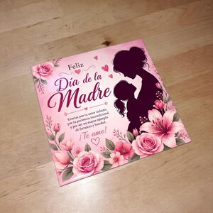 May include: A pink Mother's Day card with floral accents and the text "Feliz Día de la Madre." The card features a silhouette of a mother and child, surrounded by pink roses and lilies. The card also includes the text "¡Te amo!"