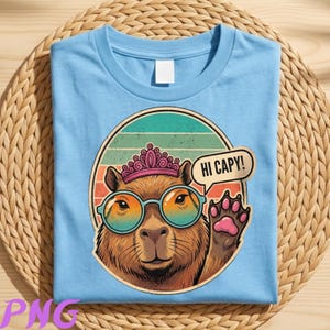 Könnte beinhalten: Hellblaues T-Shirt mit einem Cartoon-Capybara, das eine Sonnenbrille und eine Krone trägt. Das Capybara befindet sich in einem kreisförmigen Design mit einem Retro-Sonnenuntergangshintergrund. Eine Sprechblase sagt "HI CAPY!"
